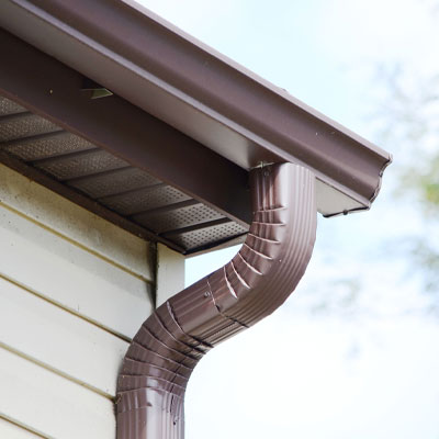 Close-up of a brown metal gutter and downspout attached to the roof of a beige panel-sided house against a blue sky background.